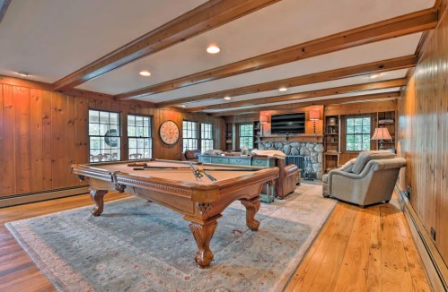 Cozy game room featuring a pool table, stone fireplace, and wooden paneling, with comfortable seating and large windows.