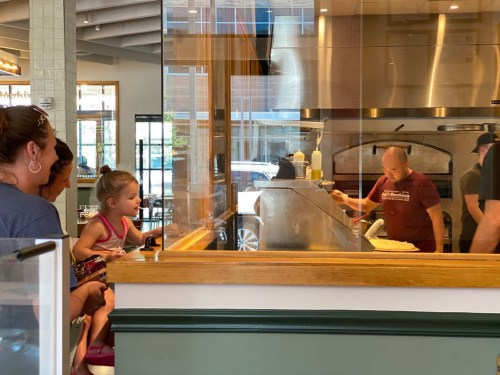A child watches a chef prepare food behind a glass counter in a casual dining setting.