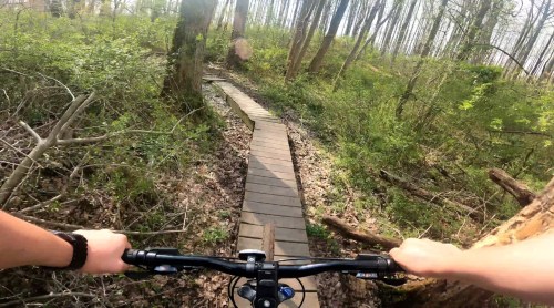 A mountain biker's view on a narrow wooden trail through a lush green forest.