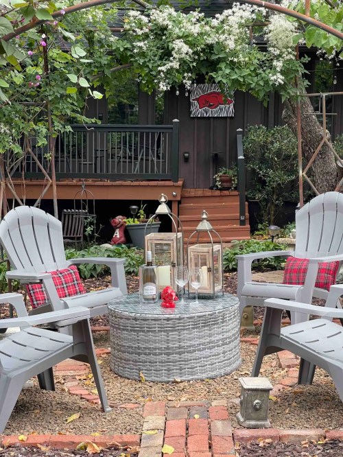 A cozy garden seating area with two chairs, a round table, lanterns, and greenery in the background.