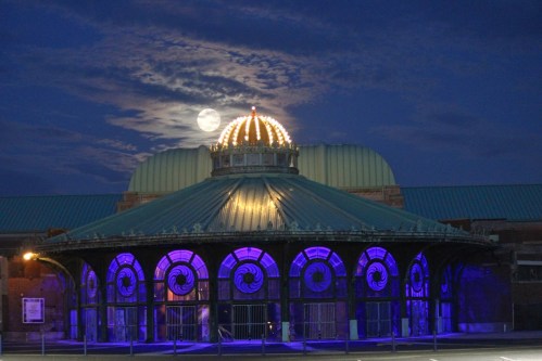 A historic building with a domed roof illuminated by purple lights, under a full moon and cloudy night sky.