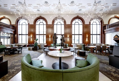 Elegant lounge with large windows, chandeliers, and green seating, featuring sculptures and a sophisticated atmosphere.