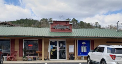 A rustic storefront with a fish sign, open sign, and a white SUV parked outside under a cloudy sky.