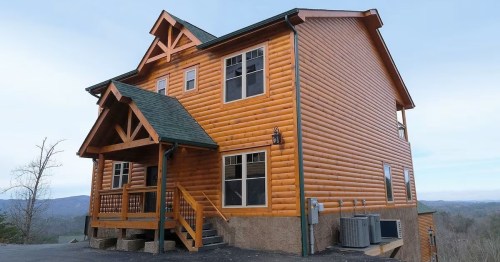 A large wooden cabin with a sloped roof, featuring a porch and multiple windows, set against a mountainous backdrop.