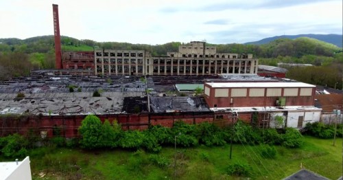 Aerial view of an abandoned factory complex surrounded by greenery and mountains in the background.