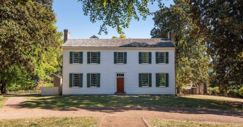 A historic white two-story house with green shutters, surrounded by trees and a clear blue sky.