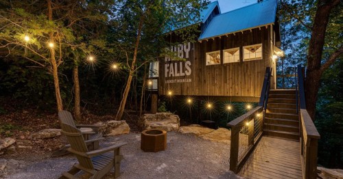 A cozy cabin at dusk, surrounded by trees, with string lights and a fire pit in front.