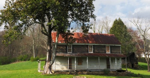 A rustic two-story house with a red metal roof, surrounded by green grass and trees under a partly cloudy sky.