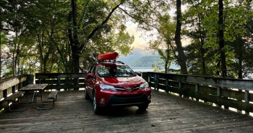 A red SUV with a kayak on top parked on a wooden deck surrounded by trees and a scenic view of a lake.