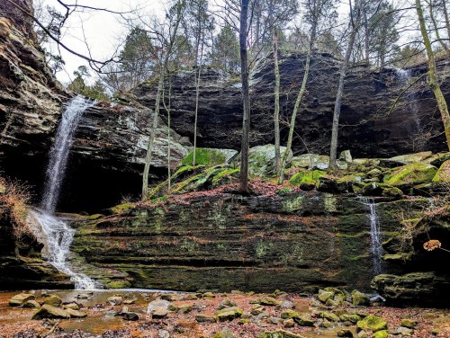 A serene landscape featuring waterfalls cascading over rocky cliffs, surrounded by trees and moss-covered stones.