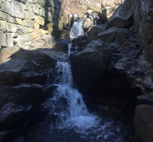 A small waterfall cascades over rocky terrain, surrounded by stone walls and natural scenery.