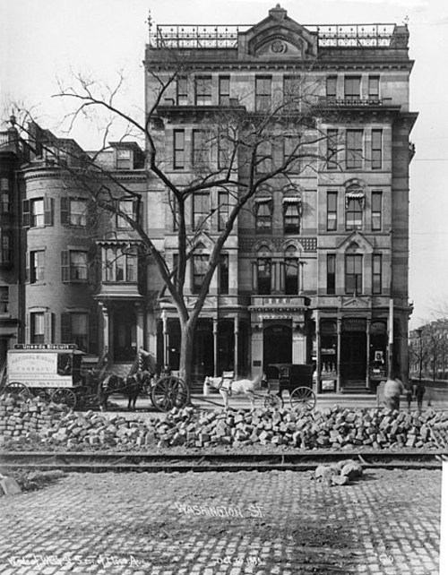 Historic street scene featuring a large building, horse-drawn carriages, and cobblestone road. Trees line the street.