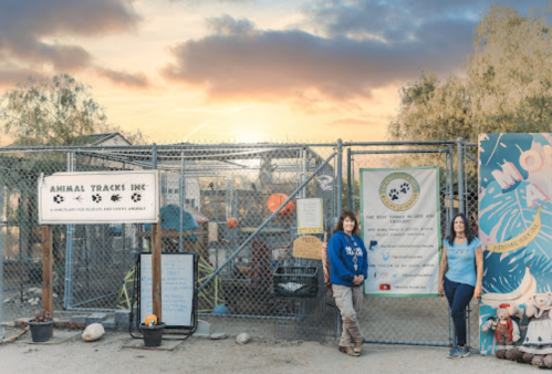 Two women stand outside a fenced animal shelter at sunset, with signs displaying the shelter's name and mission.