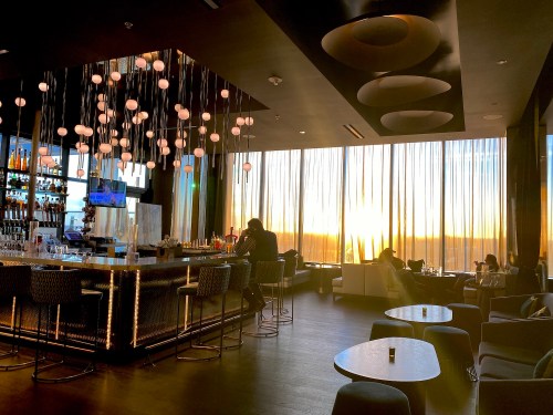 A stylish bar with modern decor, featuring a sunset view through large windows and patrons enjoying drinks.