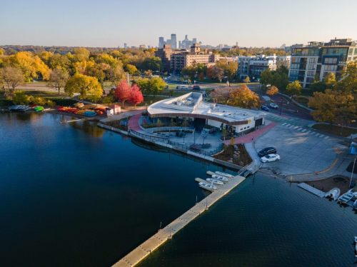 Aerial view of a lakeside park with colorful trees, a modern building, and city skyline in the background.