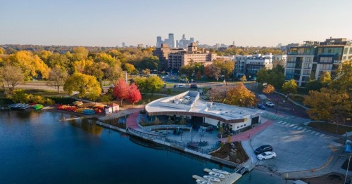 Aerial view of a lakeside area with colorful autumn trees, a modern building, and city skyline in the background.