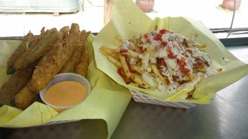 A plate of crispy fried zucchini sticks with dipping sauce next to a basket of loaded fries topped with cheese and sauce.