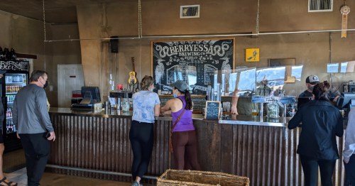 A brewery interior with customers at the counter, a chalkboard menu, and shelves of drinks in a rustic setting.