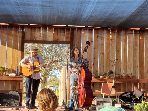 A musician plays guitar while a woman stands beside him with a double bass, performing on a wooden stage.