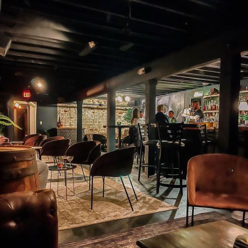 A dimly lit bar interior with wooden beams, a stone wall, and patrons seated at tables and the bar.