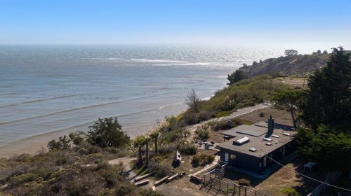 Aerial view of a coastal landscape featuring a house, beach, and calm ocean waters under a clear blue sky.