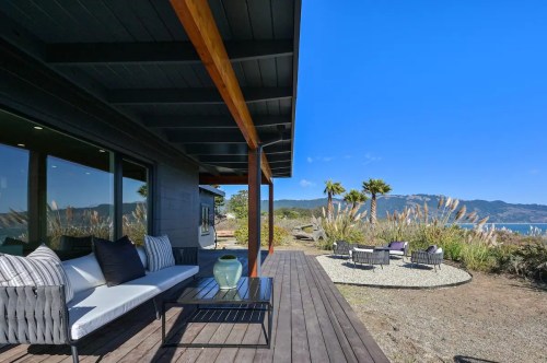 A modern patio with seating, overlooking a scenic landscape and blue sky, surrounded by greenery and palm trees.