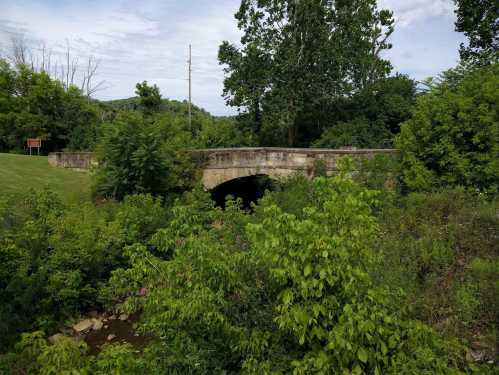 A stone bridge arches over a small creek, surrounded by lush greenery and trees under a cloudy sky.