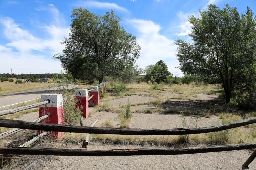 A weathered wooden fence with red and white posts, surrounded by overgrown grass and trees under a blue sky.