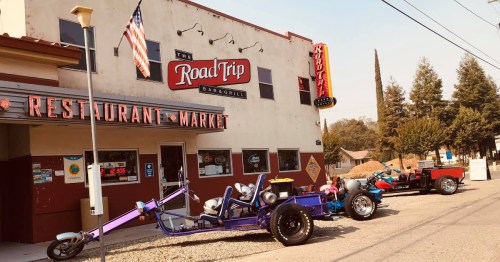 A colorful restaurant and market called "The Road Trip" with unique custom vehicles parked outside.