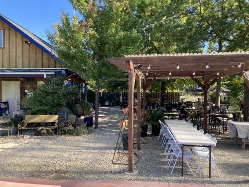 Outdoor dining area with tables under a pergola, surrounded by greenery and a rustic building in the background.