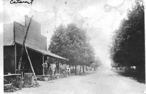 Black and white photo of a street scene with a wooden building and people standing on the porch under trees.