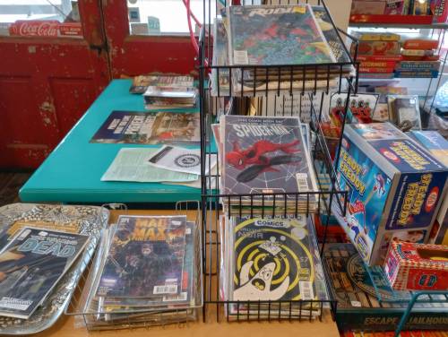 A display of comic books on a table, featuring titles like Spider-Man and The Walking Dead, with games in the background.