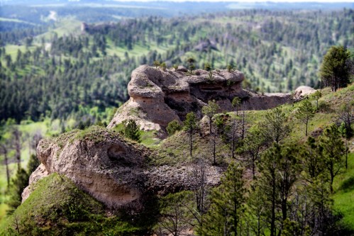A rocky outcrop surrounded by lush green hills and trees under a clear blue sky.