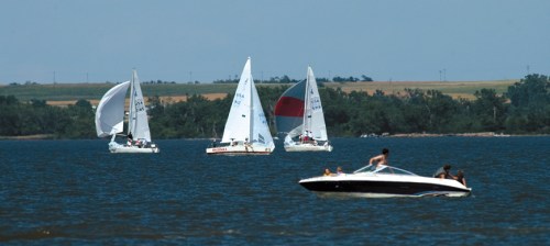 Sailboats and a motorboat on a calm lake, with green hills in the background under a clear blue sky.