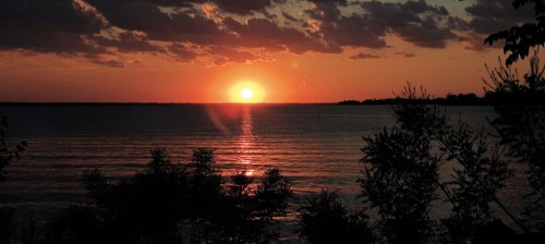 Sunset over a calm lake, with silhouettes of trees in the foreground and colorful clouds in the sky.