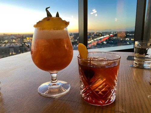 Two cocktails on a wooden table with a sunset view in the background. One is garnished with a pineapple slice.