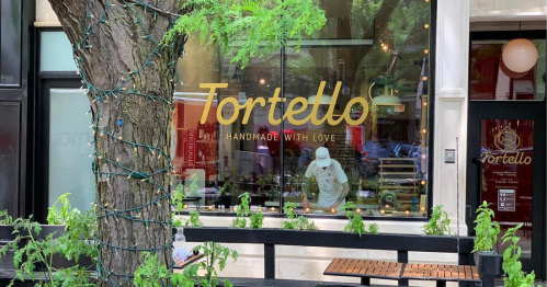 A storefront with the name "Tortello" in gold lettering, featuring a chef working inside and greenery outside.
