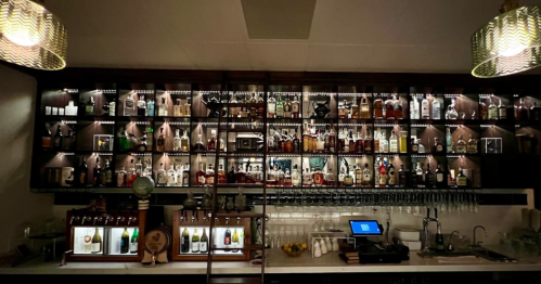 A well-stocked bar with shelves of various liquor bottles, illuminated by soft lighting and featuring a modern design.