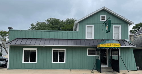 A green building with a metal roof and a yellow awning, featuring large windows and a sign hanging above the entrance.