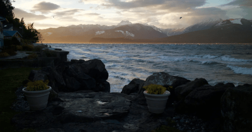 A serene lakeside view with mountains in the background, featuring rocky shores and potted plants.