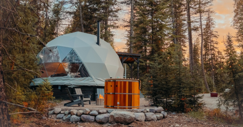 A geodesic dome cabin surrounded by trees, with a wooden hot tub and fire pit in a serene outdoor setting.