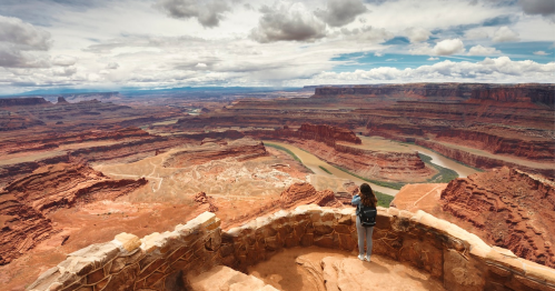 A person stands on a rocky overlook, gazing at a vast canyon landscape with winding river and cloudy sky.