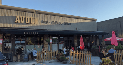 Exterior of Avol, a restaurant with outdoor seating, umbrellas, and a wooden fence, under a clear blue sky.