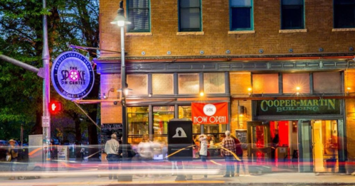 A lively street scene at night featuring a restaurant with a "Now Open" sign and the Cooper-Martin Building nearby.