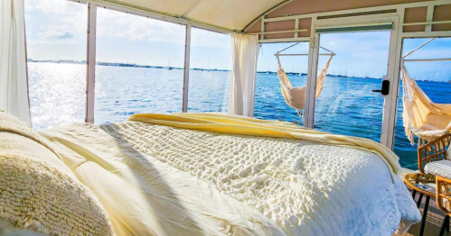 Cozy bedroom with a view of water, featuring a bed, soft blankets, and a hammock by large windows.