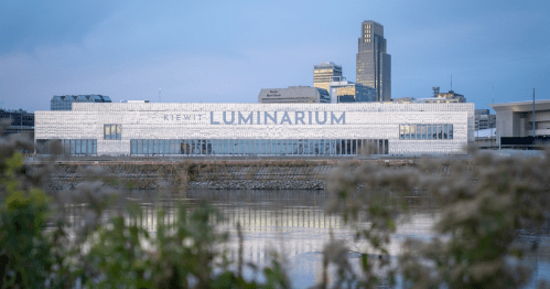 A modern building labeled "Luminarium" reflects in a river, with city skyline and greenery in the foreground.