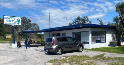 A small café with a blue roof and sign, featuring a parked car and outdoor seating under a clear sky.