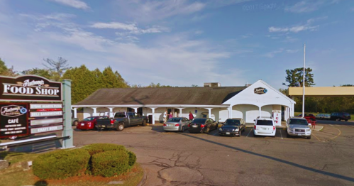 A food shop with a white exterior, surrounded by parked cars and trees, on a sunny day.