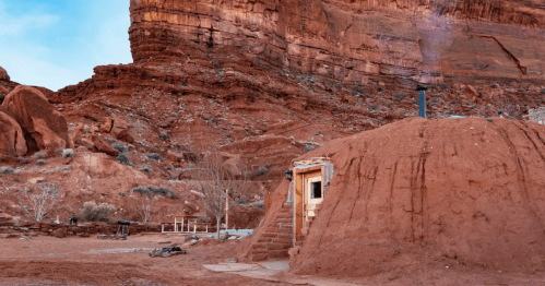 A small earthen structure nestled against red rock formations, with a chimney and steps leading up to the entrance.