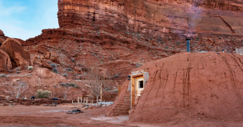 A small earthen structure nestled against red rock formations, with a chimney and steps leading up to the entrance.